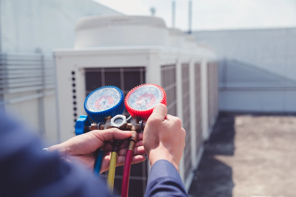 Technicien HVAC tenant un manifold de mesure de pression avec manomètres haute et basse pression, jaunes et rouges, devant une unité de climatisation extérieure résidentielle