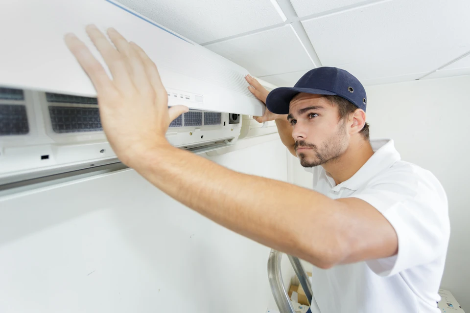 Technicien en casquette bleue et polo blanc effectue la maintenance d'une unité de climatisation murale dans un bureau aux plafonds blancs